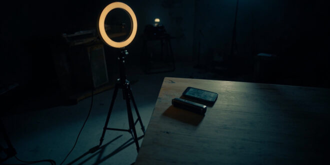 Dark recording studio with inactive ring light, smartphone on desk, microphone stand casting long shadow in empty room