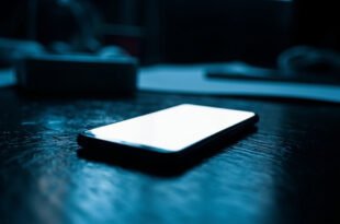 Illuminated smartphone on dark wooden desk surface with dramatic shadows and cold blue atmospheric lighting