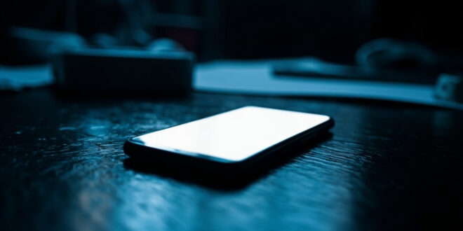 Illuminated smartphone on dark wooden desk surface with dramatic shadows and cold blue atmospheric lighting
