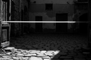 Weathered wooden door ajar, empty cobblestone courtyard, forensic tape across entrance, abandoned rural house exterior