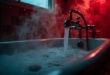 Empty bathtub with steam rising from scalding water, chrome faucet in sharp focus against dark tiles