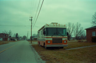 Large weathered Winnebago RV parked on residential street beside redbrick house under gray overcast sky