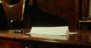 A folded white note with black pen resting on dark wooden desk surface in formal office setting