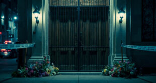 Closed synagogue doors with security barriers, police tape, and flower tributes on wet pavement under emergency lighting