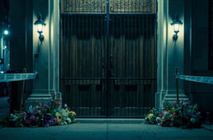 Closed synagogue doors with security barriers, police tape, and flower tributes on wet pavement under emergency lighting