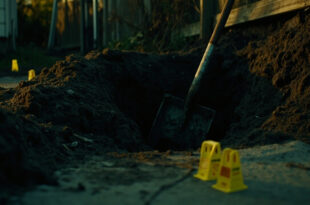 Shovel in dark soil next to yellow evidence markers on rural property with forensic equipment scattered around