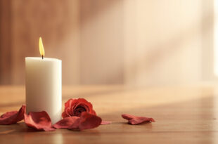 White candle flame glowing softly among scattered pink rose petals on weathered wood surface