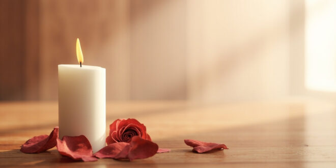 White candle flame glowing softly among scattered pink rose petals on weathered wood surface