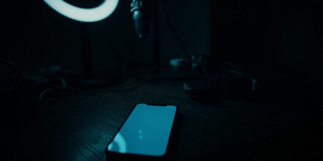 Dark smartphone on wooden desk beside silent microphone and dimmed ring light in shadowy room