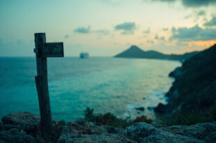 Solitary wooden trail marker on cliff edge above turquoise coral reef waters under overcast sky