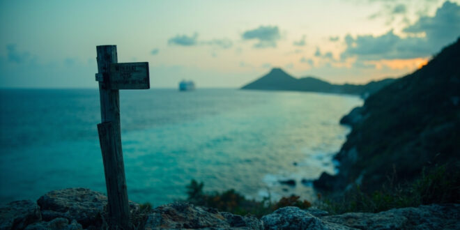 Solitary wooden trail marker on cliff edge above turquoise coral reef waters under overcast sky