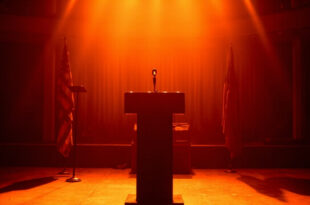 Empty podium with microphone under spotlight on stage with American flags and dark blue curtains in background