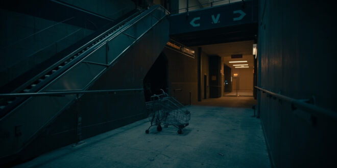 Empty shopping center escalator descending toward dim exit, abandoned cart near emergency door, cold blue lighting