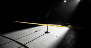 Empty theater stage with single microphone stand under spotlight, police barrier tape visible in foreground