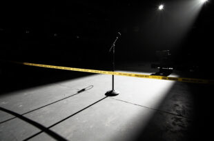 Empty theater stage with single microphone stand under spotlight, police barrier tape visible in foreground