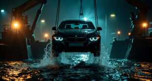 Black sedan dripping water suspended by crane cable above dark canal surface with emergency lights reflecting